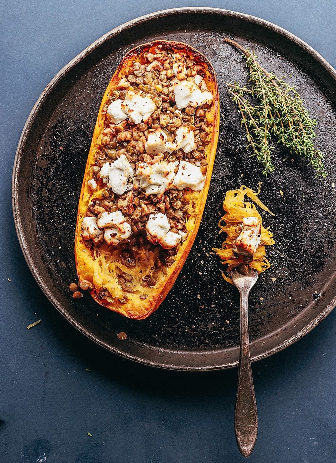 Ein gebratener halber Spaghettikürbis, gefüllt mit Linsen und überbacken mit geschmolzenem Käse, liegt auf einem runden Backblech, daneben frischer Thymian. Auf dem Tablett liegt eine Gabel, auf der ein Bissen des Gerichts liegt.