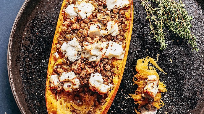 Ein gebratener halber Spaghettikürbis, gefüllt mit Linsen und überbacken mit geschmolzenem Käse, liegt auf einem runden Backblech, daneben frischer Thymian. Auf dem Tablett liegt eine Gabel, auf der ein Bissen des Gerichts liegt.