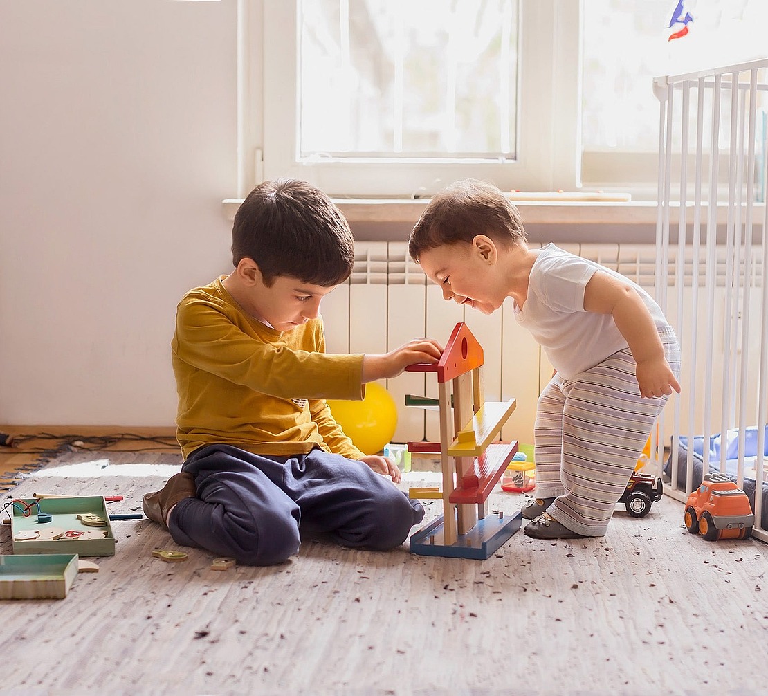 Zwei kleine Kinder spielen zusammen auf einem Teppich in einem sonnenbeschienenen Raum mit einem bunten Holzspielzeug. Ein Kind sitzt konzentriert, während das andere steht und sich neugierig anlehnt. Spielzeug und Bücher sind um sie herum verstreut.