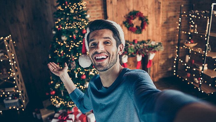 Ein lächelnder Mann mit Weihnachtsmannmütze macht ein Selfie in einem festlich geschmückten Raum mit einem Weihnachtsbaum, Kranz, Girlanden, Lichtern und eingepackten Geschenken.