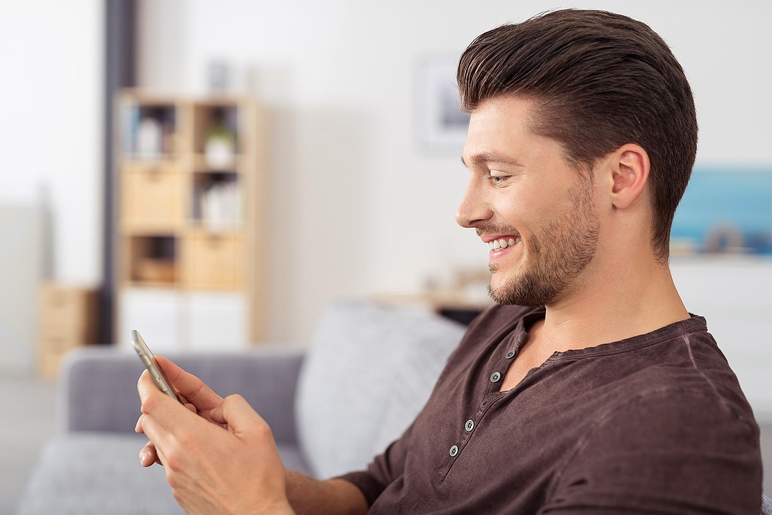 Ein junger Mann mit kurzen braunen Haaren und einem gestutzten Bart sitzt lächelnd auf einer Couch und schaut auf sein Smartphone. Der Hintergrund zeigt einen verschwommenen Raum mit Regalen und einem Gemälde.