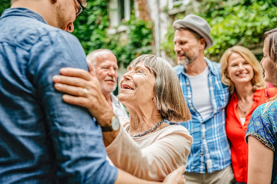 Eine lächelnde ältere Frau umarmt einen jüngeren Mann im Freien, umgeben von lächelnden Erwachsenen. Die Gruppe steht eng beieinander, wirkt glücklich und genießt die Gesellschaft des anderen in einer üppigen, grünen Umgebung.