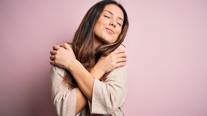 Eine junge Frau mit langen braunen Haaren umarmt sich selbst und lächelt mit geschlossenen Augen, während sie vor einem rosa Hintergrund steht. Sie sieht zufrieden und entspannt aus und trägt einen hellen Pullover.