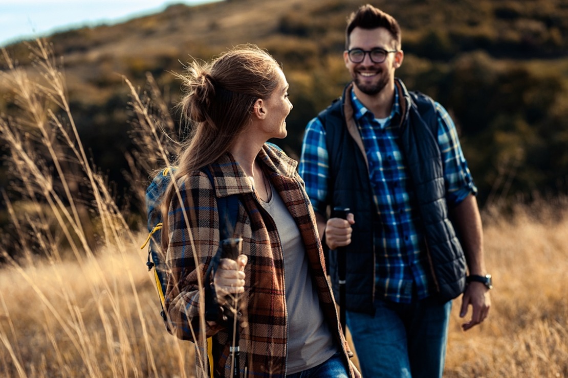 Ein lächelnder Mann und eine lächelnde Frau in legerer Outdoor-Kleidung wandern an einem sonnigen Tag durch ein grasbewachsenes Feld, halten Trekkingstöcke und schauen sich an. Im Hintergrund sind sanfte Hügel und Bäume zu sehen.