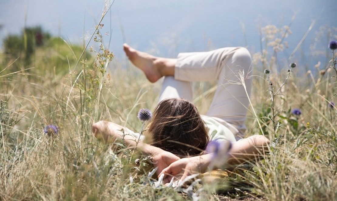 Eine Person liegt auf dem Rücken im hohen Gras, die Arme hinter dem Kopf und die Beine entspannt, umgeben von Wildblumen an einem sonnigen Tag, mit Wasser im verschwommenen Hintergrund.