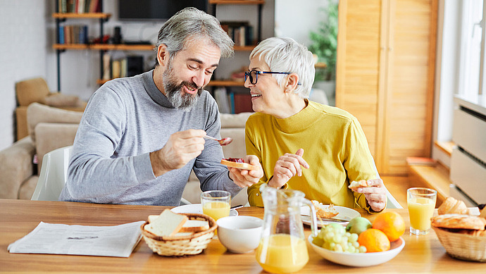 Ein älteres Paar sitzt an einem Tisch und genießt gemeinsam das Frühstück. Sie lächeln und unterhalten sich. Der Tisch ist mit Orangensaft, Obst, Brot und Gebäck gedeckt. Eine Zeitung liegt daneben, und die Atmosphäre ist gemütlich und sonnig.