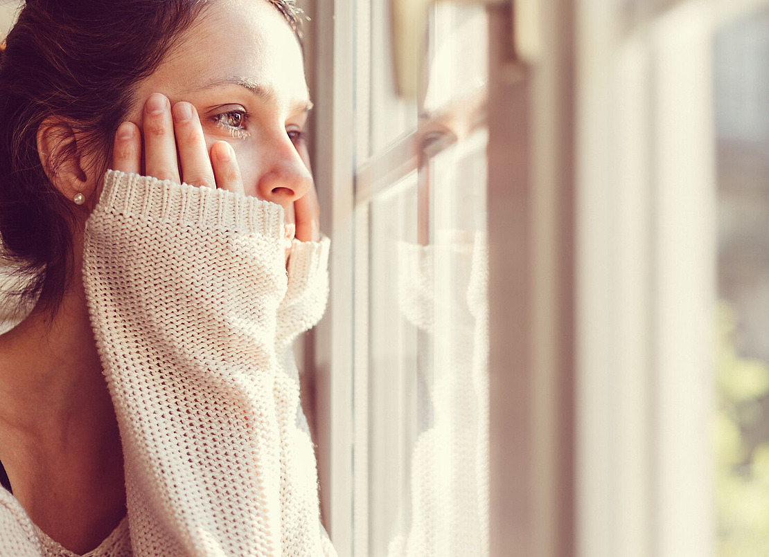 Eine Frau blickt nachdenklich aus dem Fenster. Ihre Hände ruhen sanft auf ihren Wangen. Sie trägt einen kuscheligen, hellen Pullover und natürliches Licht erhellt ihr Gesicht sanft. Der Hintergrund ist unscharf und suggeriert eine ruhige Szene im Freien.