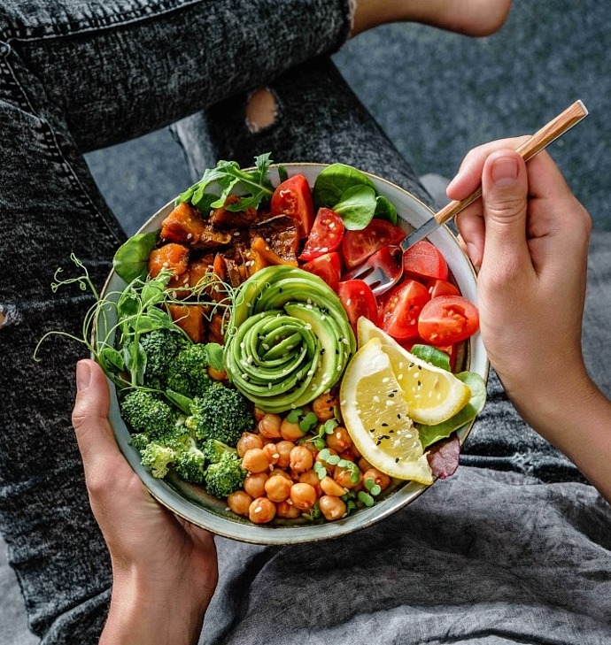 Eine Person in zerrissenen schwarzen Jeans hält eine bunte Schüssel mit Tomaten, Avocado-Rose, Zitrone, Kichererbsen, Brokkoli, Grünzeug und gebratenem Gemüse und isst im Schneidersitz mit einer Gabel.