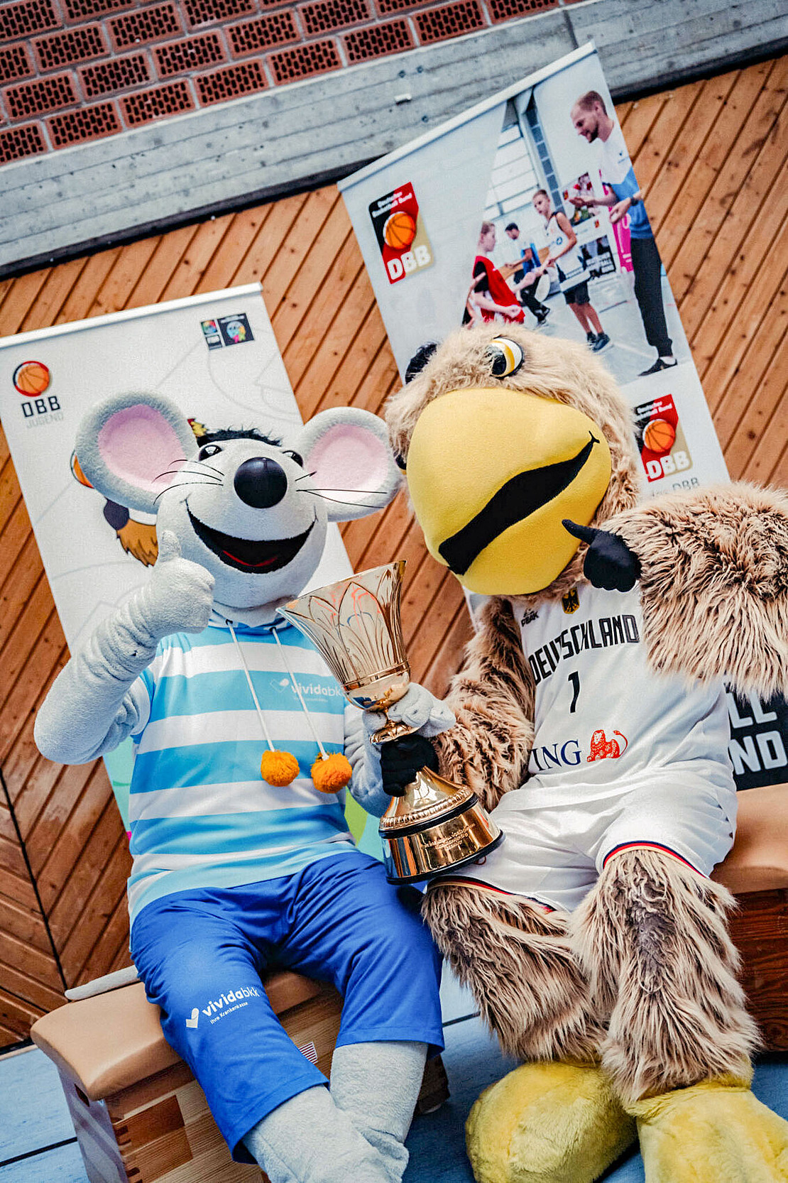 Zwei Sportmaskottchen – eine lächelnde Maus in gestreiftem Hemd und Hose und ein großer Vogel in einem Basketballtrikot – sitzen nebeneinander und halten jeweils eine Trophäe in der Hand. Im Hintergrund sind Veranstaltungsbanner zu sehen.