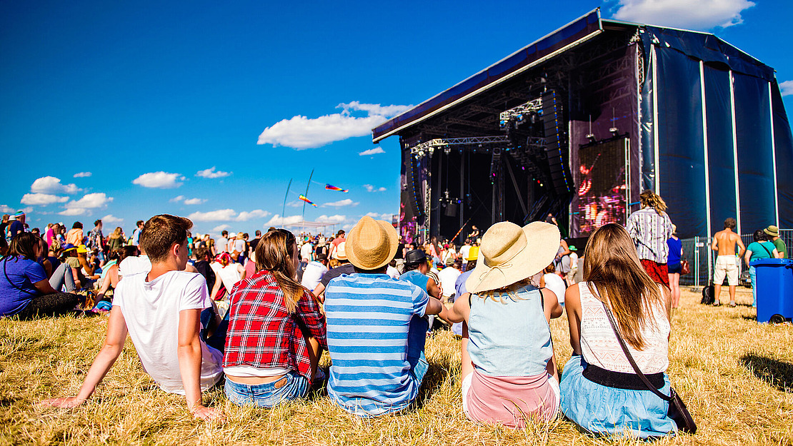 Eine Gruppe von Menschen sitzt auf einer Wiese vor einer Freilichtbühne bei einem Musikfestival. Im Hintergrund sind eine Menschenmenge, bunte Fahnen und ein strahlend blauer Himmel zu sehen. Einige tragen Hüte und sommerliche Kleidung.