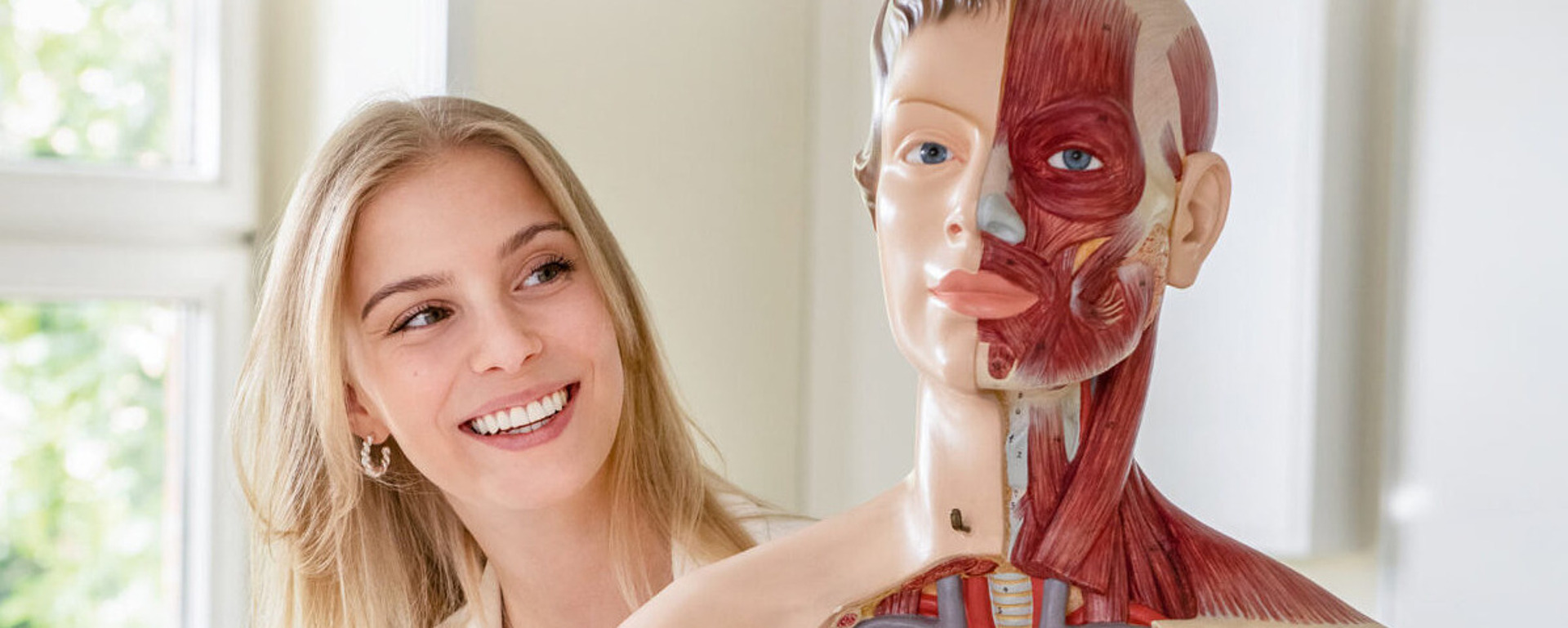 Eine lächelnde Frau steht neben einem anatomischen menschlichen Torsomodell, das Muskeln und innere Organe zeigt, in einem hellen Klassenzimmer mit Schreibtischen und Fenstern im Hintergrund.