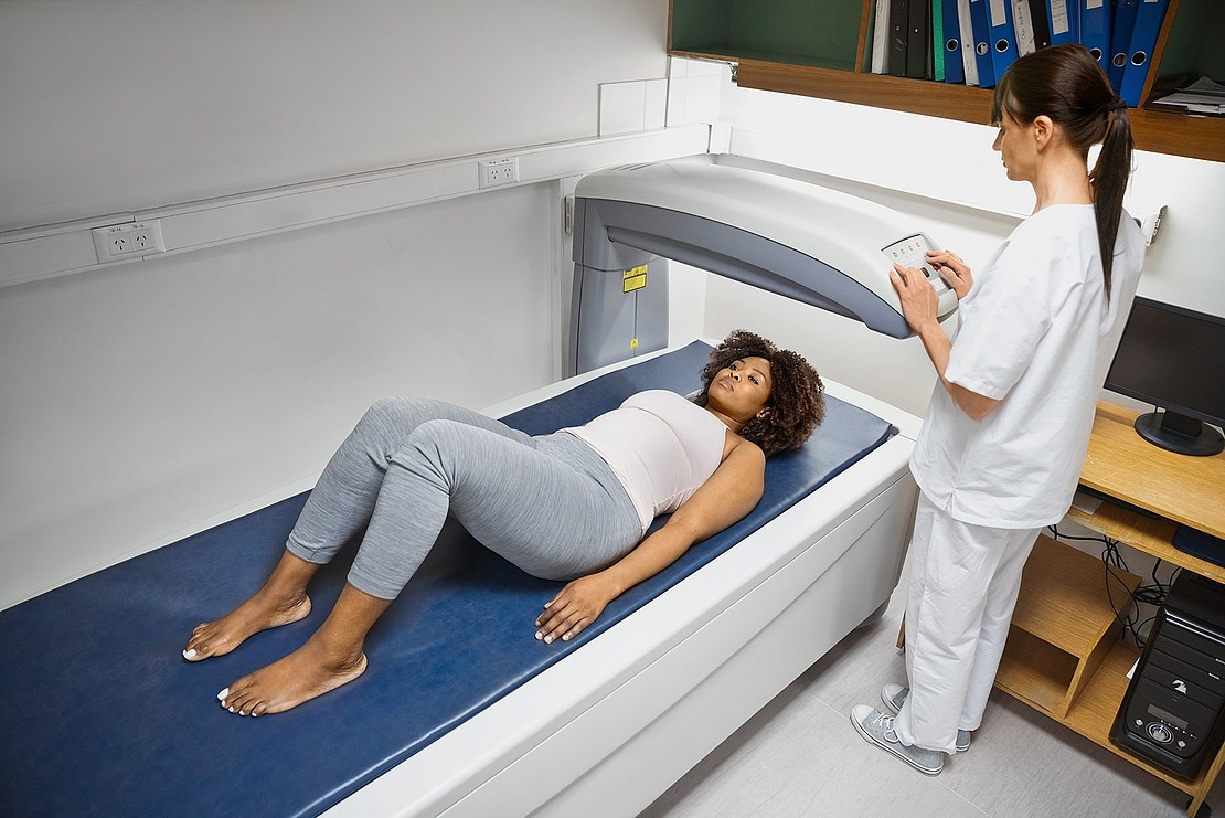 Eine Frau liegt auf dem Rücken auf einem medizinischen Scannertisch, während eine medizinische Fachkraft ein Bedienfeld bedient und einen diagnostischen Scan in einem klinischen Raum durchführt.