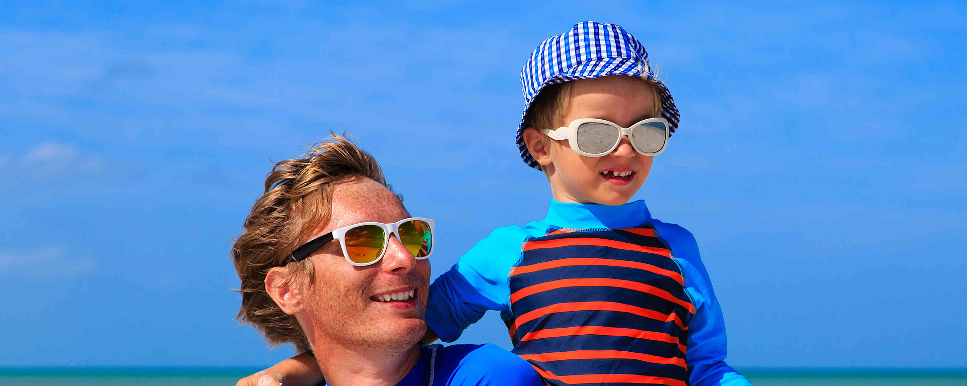 Ein Mann und ein Kind mit Sonnenbrille, Hut und Badeshirt lächeln zusammen an einem sonnigen Strand mit blauem Himmel und ruhigem Meer im Hintergrund.