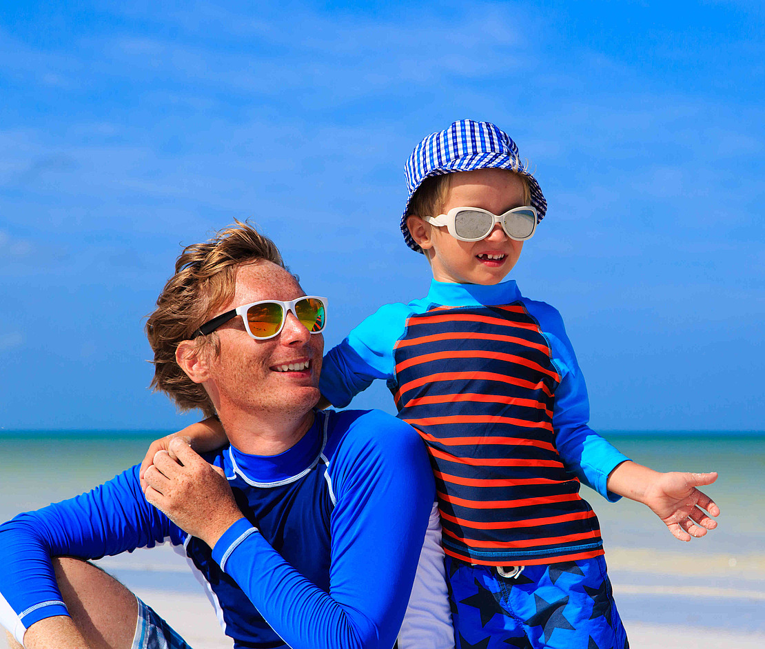 Ein Mann und ein Kind mit Sonnenbrille, Hut und Badeshirt lächeln zusammen an einem sonnigen Strand mit blauem Himmel und ruhigem Meer im Hintergrund.