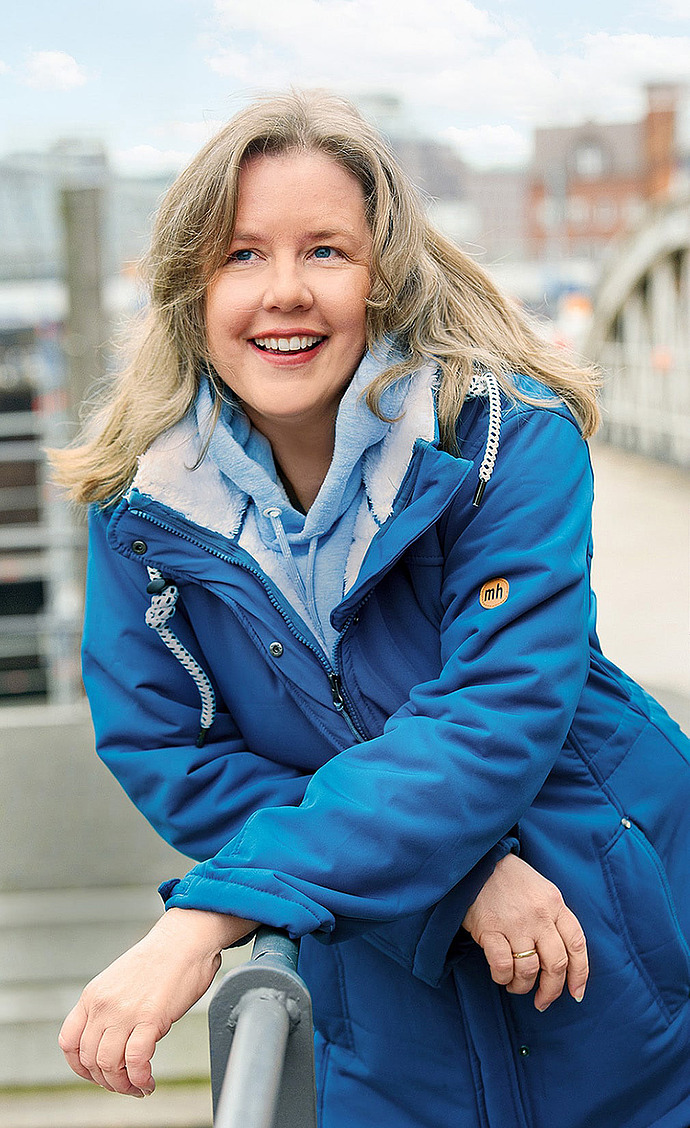 Eine lächelnde Frau mit blondem Haar, die einen blauen Wintermantel und einen hellblauen Kapuzenpulli trägt, lehnt sich an ein Metallgeländer im Freien mit einem unscharfen städtischen Hintergrund.
