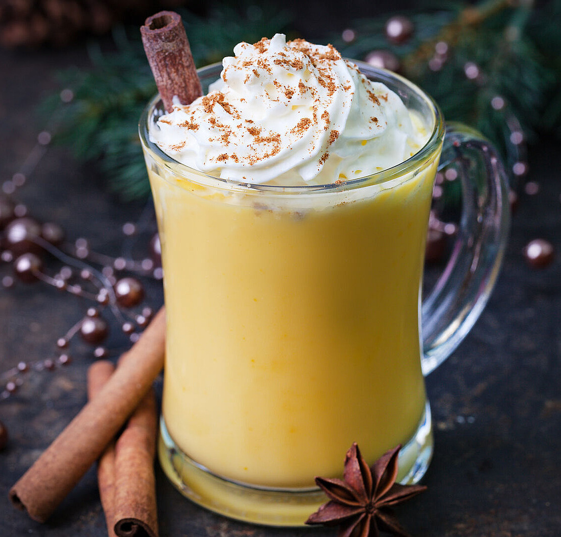 Ein Glasbecher mit Eierlikör, gekrönt mit Schlagsahne und einer Prise Gewürzen, garniert mit einer Zimtstange. Ganze Zimtstangen und Sternanis stehen neben dem Becher, mit festlichem Grün im Hintergrund.