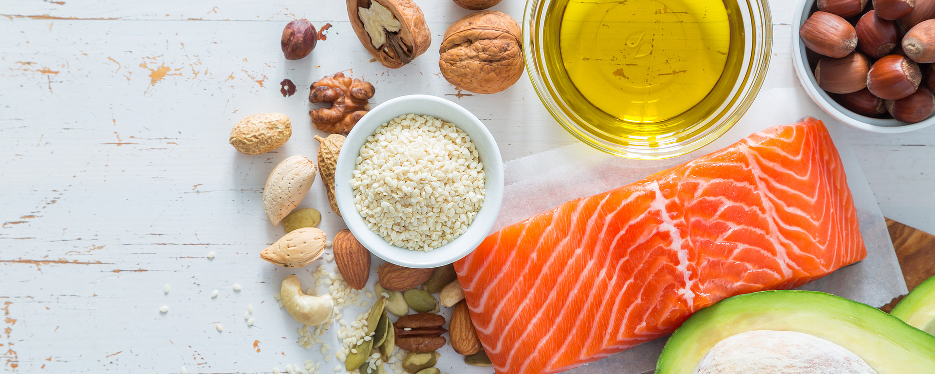 Auf einem weißen, rustikalen Tisch stehen verschiedene gesunde Lebensmittel, die reich an guten Fetten sind, darunter roher Lachs, eine halbe Avocado, gemischte Nüsse, Samen und eine Schale mit Olivenöl.