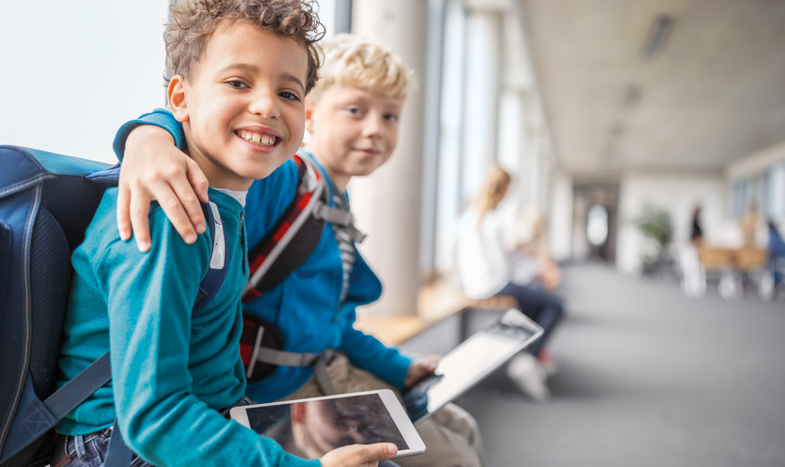 Zwei Jungen mit Rucksäcken sitzen auf einer Bank in einem hellen Flur und lächeln in die Kamera. Ein Junge hat seinen Arm um den anderen gelegt, und beide halten Tablet-Geräte in der Hand. Im Hintergrund sitzen weitere Personen.