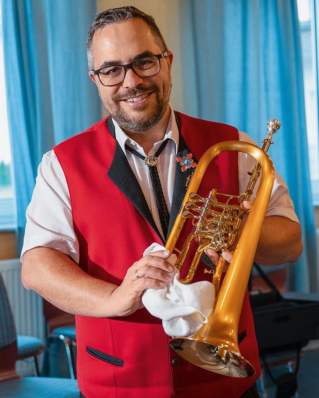 Ein lächelnder Mann mit Brille und roter Weste steht in einem Raum mit blauen Vorhängen und hält ein Blechblasinstrument und einen Putzlappen in der Hand. Im Hintergrund sind leere Stühle und Musikgeräte zu sehen.