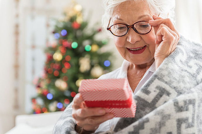 Eine ältere Frau lächelt, während sie ein kleines, eingepacktes Geschenk öffnet. Sie trägt eine Brille und eine gemusterte Decke. Ein geschmückter Weihnachtsbaum mit bunten Lichtern ist unscharf im Hintergrund zu sehen.