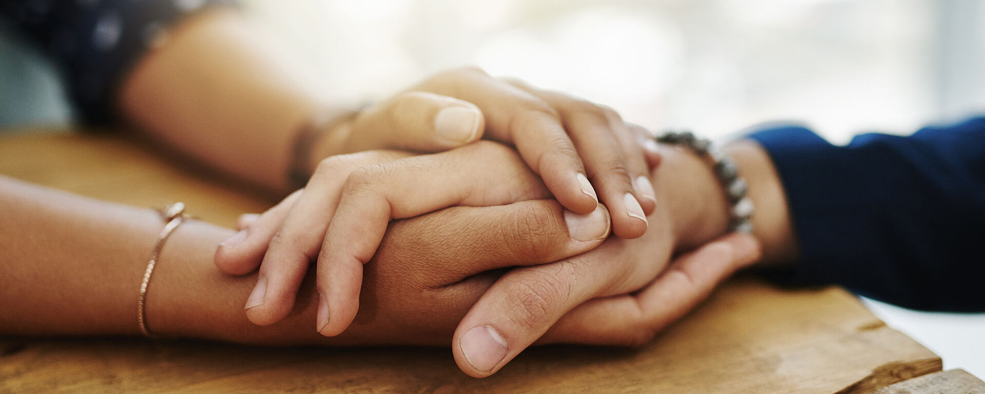 Zwei Menschen sitzen an einem Holztisch und halten einander sanft die Hände in einer tröstenden und unterstützenden Geste. Der Fokus liegt auf ihren Händen, mit sanftem Sonnenlicht im Hintergrund.