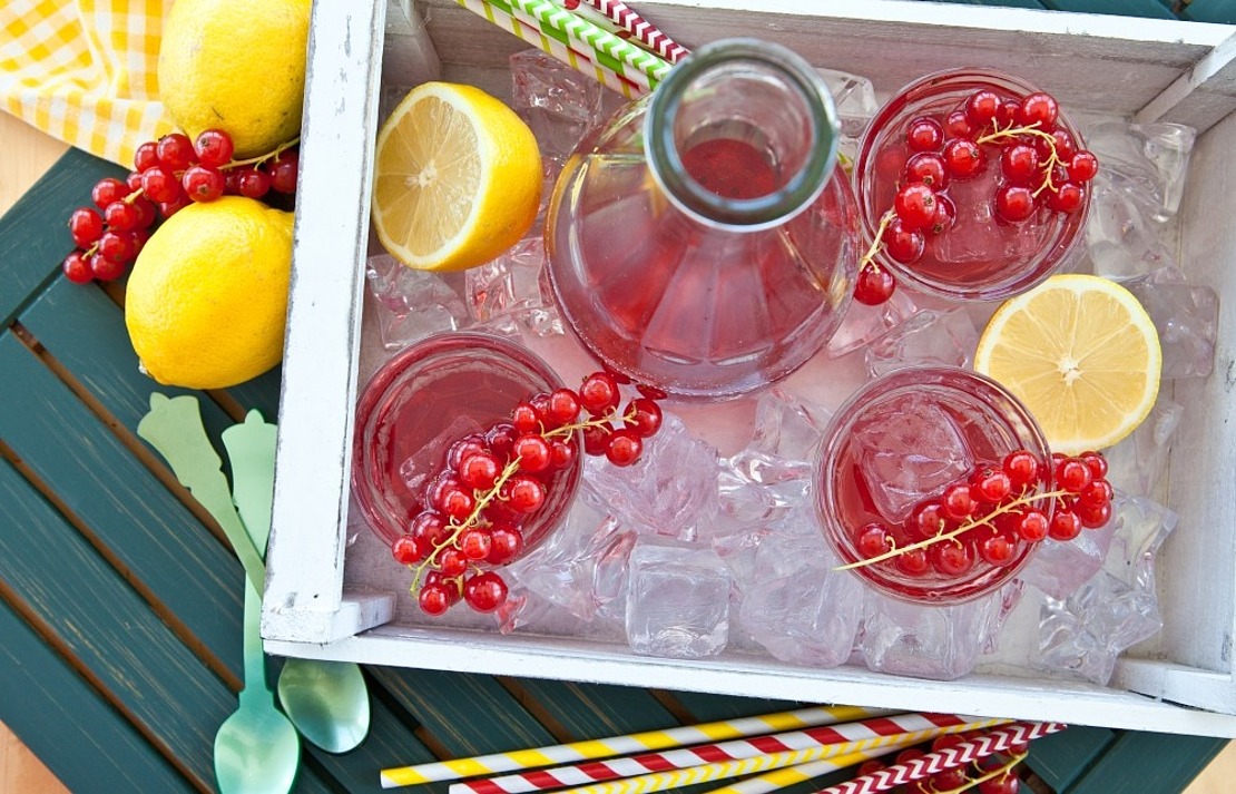 Ein weißes Tablett, gefüllt mit Eis, eine Glaskaraffe und zwei Gläser mit rotem Getränk, garniert mit roten Johannisbeeren und Zitronenscheiben. Zitronen, bunte Strohhalme und Löffel sind in der Nähe auf einem grünen Tisch angeordnet.