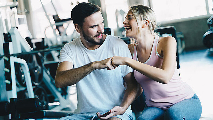 Ein Mann und eine Frau sitzen in einem Fitnessstudio, lächeln und stoßen mit den Fäusten aneinander. Im Hintergrund sind Fitnessgeräte zu sehen. Beide wirken nach dem Training glücklich und entspannt.
