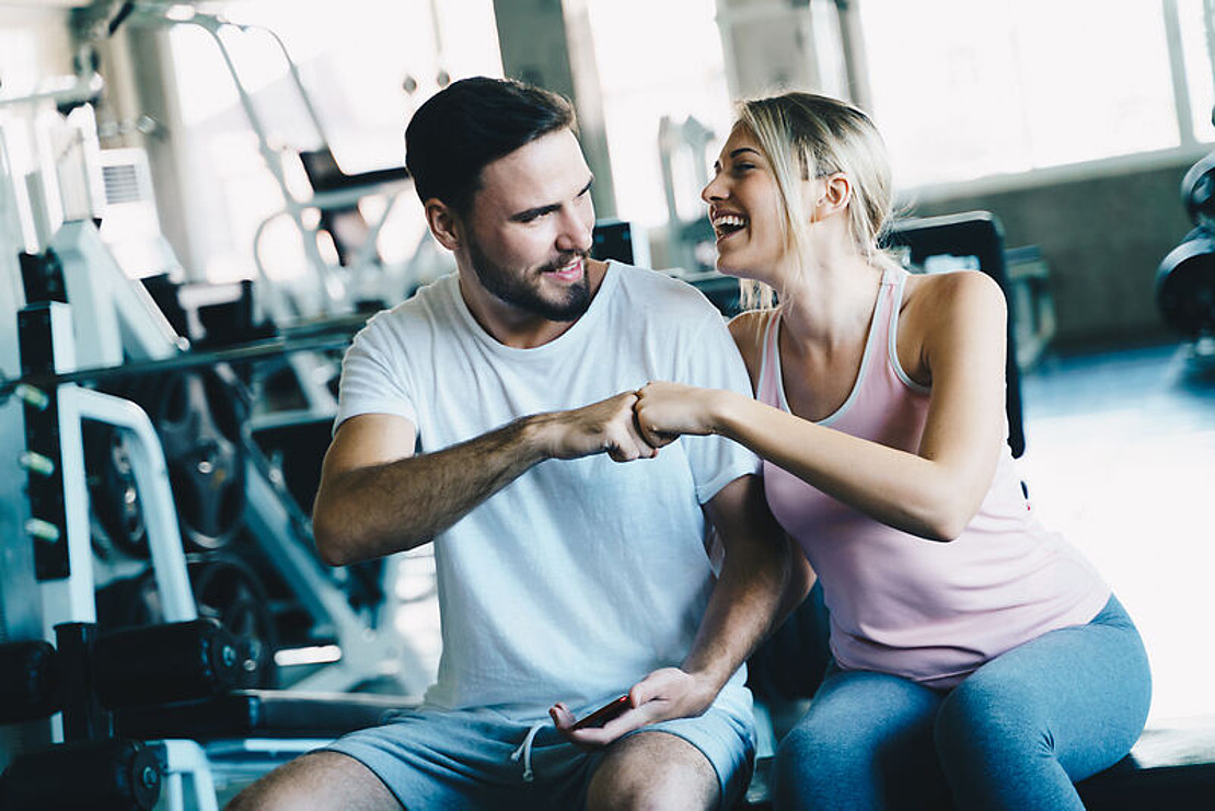Ein Mann und eine Frau sitzen in einem Fitnessstudio, lächeln und stoßen mit den Fäusten aneinander. Im Hintergrund sind Fitnessgeräte zu sehen. Beide wirken nach dem Training glücklich und entspannt.