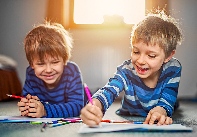 Zwei Jungen in gestreiften Hemden liegen auf dem Boden, lächeln und zeichnen mit Buntstiften auf Papier, während hinter ihnen das Sonnenlicht durch ein Fenster fällt.