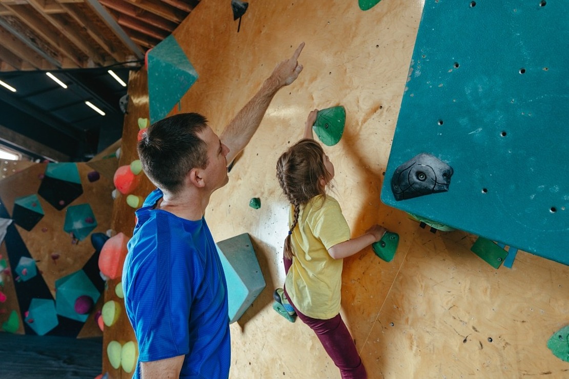 Ein Mann in einem blauen Hemd weist ein junges Mädchen in einem gelben Hemd an, an einer Indoor-Kletterwand zu klettern. Die Wand ist voller bunter Griffe und geometrischer Formen, und das Mädchen wirkt konzentriert und mit vollem Einsatz bei der Aktivität.