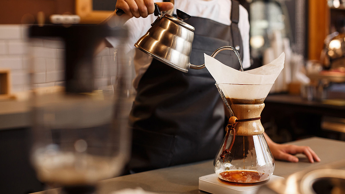 Ein Barista in einer dunklen Schürze gießt heißes Wasser aus einem Schwanenhalskessel in eine Chemex-Kaffeemaschine mit Papierfilter und bereitet an einer Café-Theke Pour-Over-Kaffee zu.