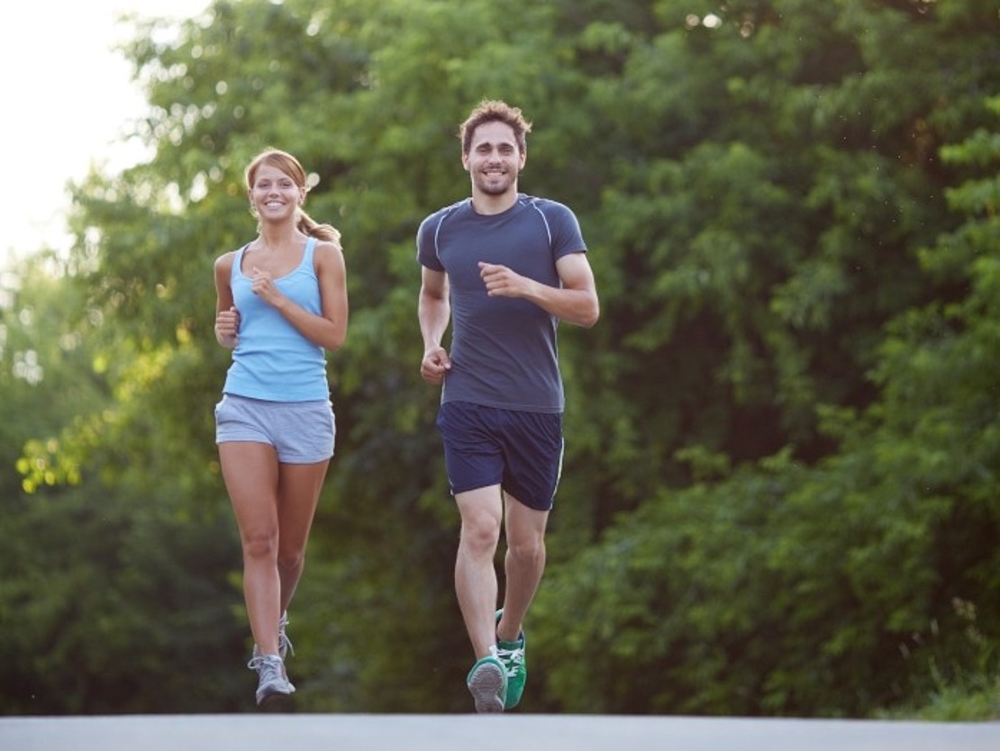 Ein Mann und eine Frau joggen im Freien auf einem gepflasterten Weg mit grünen Bäumen im Hintergrund, beide lächeln und tragen sportliche Kleidung an einem sonnigen Tag.