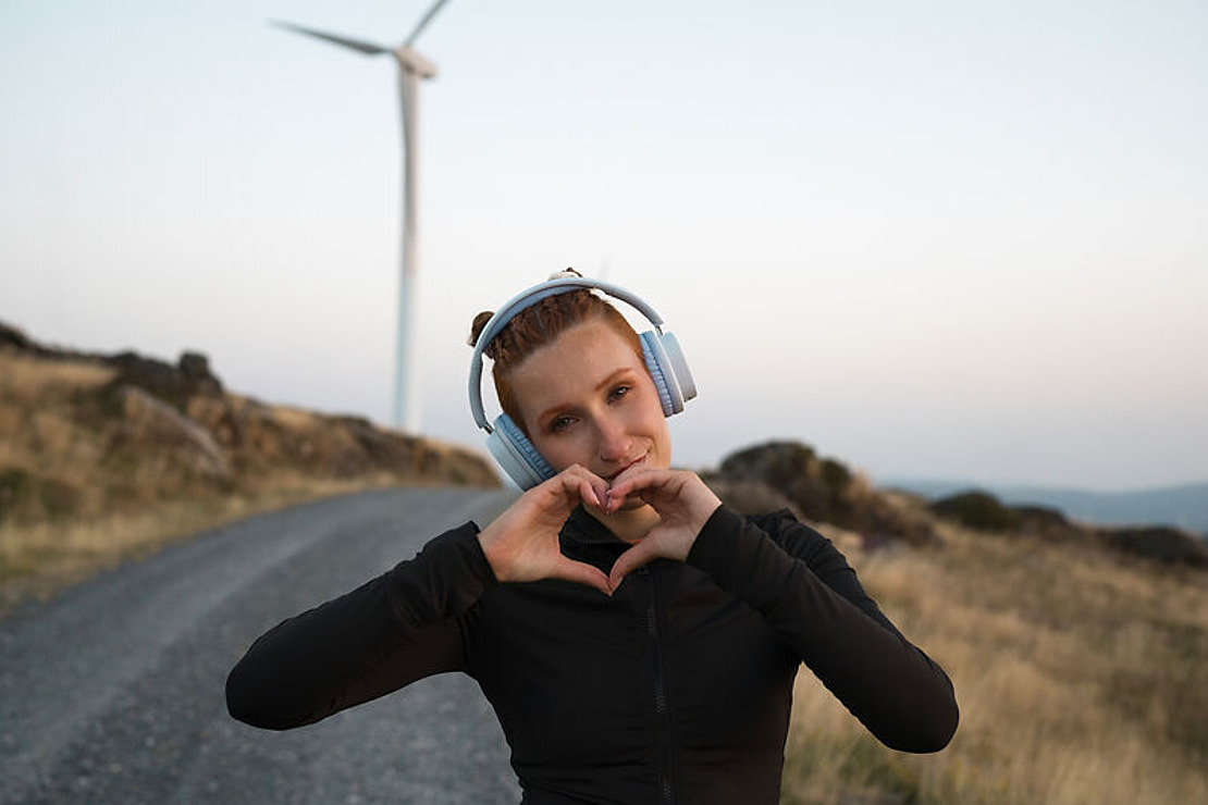 Eine junge Frau mit Kopfhörern formt mit ihren Händen eine Herzform, während sie auf einer Schotterstraße in der Nähe einer Windkraftanlage in einer ländlichen Landschaft bei Sonnenuntergang steht.