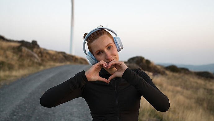 Eine junge Frau mit Kopfhörern formt mit ihren Händen eine Herzform, während sie auf einer Schotterstraße in der Nähe einer Windkraftanlage in einer ländlichen Landschaft bei Sonnenuntergang steht.