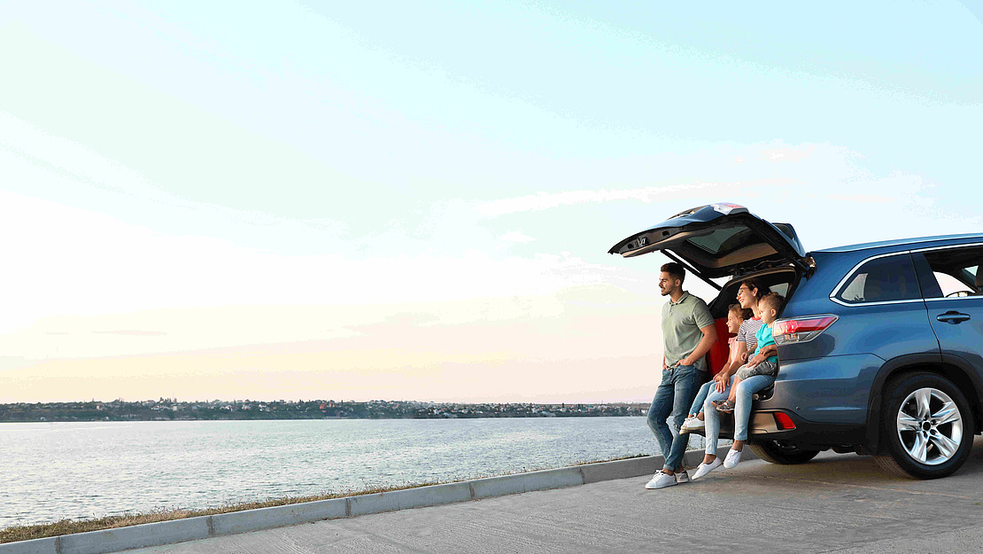 Eine vierköpfige Familie sitzt im offenen Kofferraum eines blauen Geländewagens, der am Wasser geparkt ist, und genießt die Aussicht auf den Sonnenuntergang. Die Eltern sitzen am Rand, mit zwei Kindern zwischen ihnen, lächelnd und entspannt.