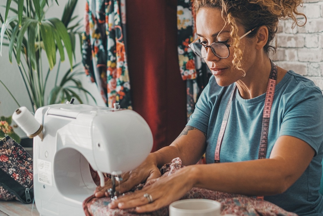 Eine Frau mit lockigem Haar und Brille arbeitet an einer Nähmaschine und konzentriert sich auf den Stoff. Sie trägt ein blaues Hemd und ein Maßband um den Hals. Im Hintergrund sind bunte Stoffe und eine Topfpflanze zu sehen.