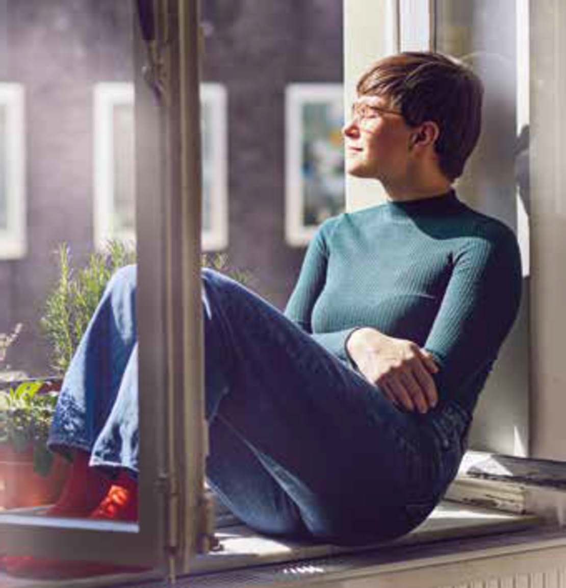 Eine Person mit kurzen Haaren und Brille sitzt auf einer Fensterbank, schaut nach draußen und genießt das Sonnenlicht. Sie trägt ein grünes, langärmeliges Oberteil, blaue Jeans und rote Socken und hat Pflanzen in der Nähe auf der Fensterbank stehen.