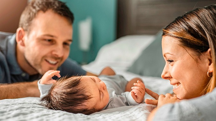 Ein lächelndes Paar liegt auf einem Bett und betrachtet liebevoll sein neugeborenes Baby, das zwischen ihnen liegt und seine kleinen Hände nach oben streckt. Die Szene ist warm und intim und unterstreicht die familiäre Verbundenheit.