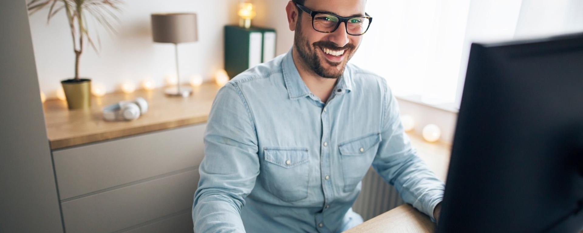 Ein bärtiger Mann mit Brille und hellblauem Hemd lächelt, während er einen Computer an einem Schreibtisch in einem hellen, modernen Heimbüro mit Regalen und dekorativen Lichtern im Hintergrund benutzt.