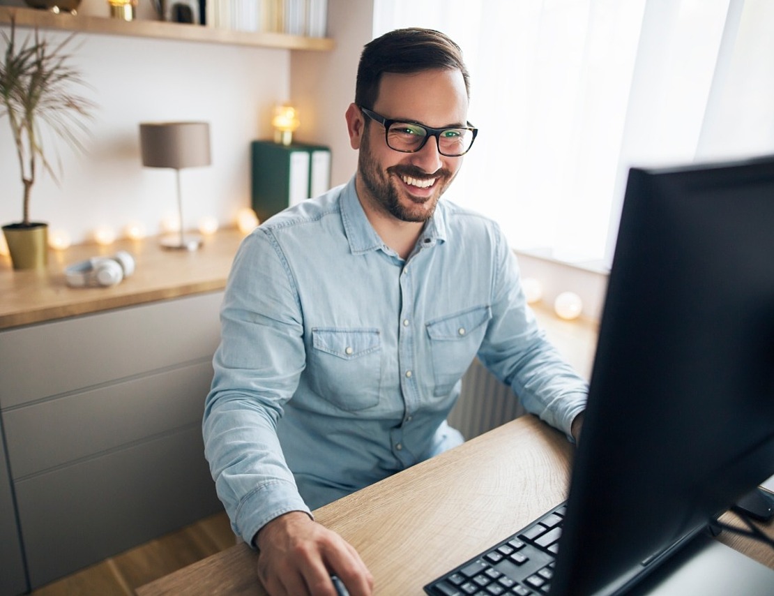 Ein bärtiger Mann mit Brille und hellblauem Hemd lächelt, während er einen Computer an einem Schreibtisch in einem hellen, modernen Heimbüro mit Regalen und dekorativen Lichtern im Hintergrund benutzt.