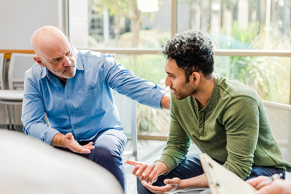 Ein Mann mittleren Alters tröstet einen jüngeren Mann sanft während eines ernsten Gesprächs in einem hellen Raum mit großen Fenstern. Der jüngere Mann sieht verzweifelt aus, und der ältere Mann legt ihm unterstützend die Hand auf die Schulter.