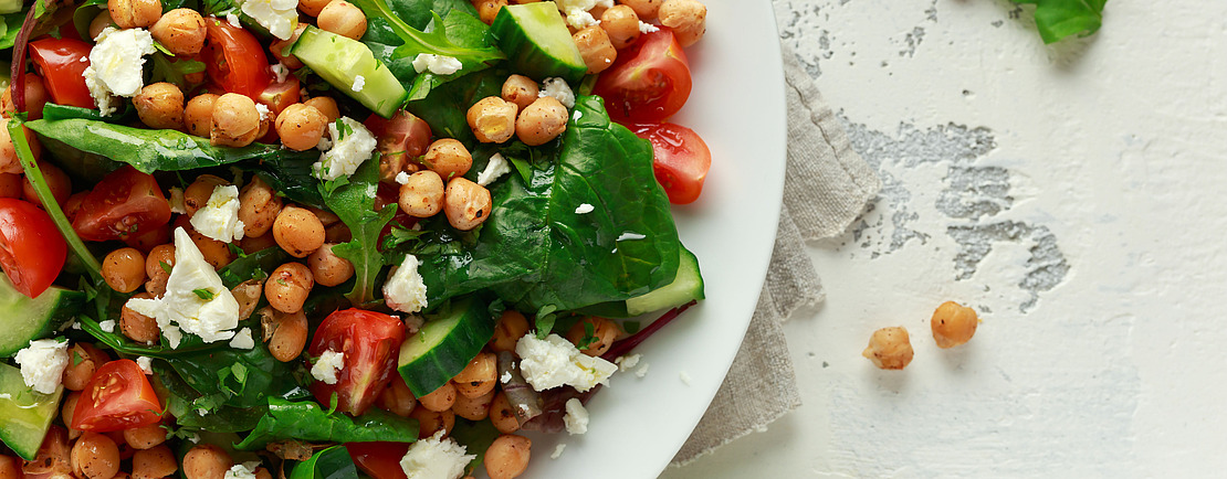 Ein frischer Salat mit Spinat, Kirschtomaten, Gurken, Kichererbsen und Feta auf einem weißen Teller, garniert mit einer Zitronenspalte. In der rechten oberen Ecke erscheint ein "MACH ES!"-Schild.