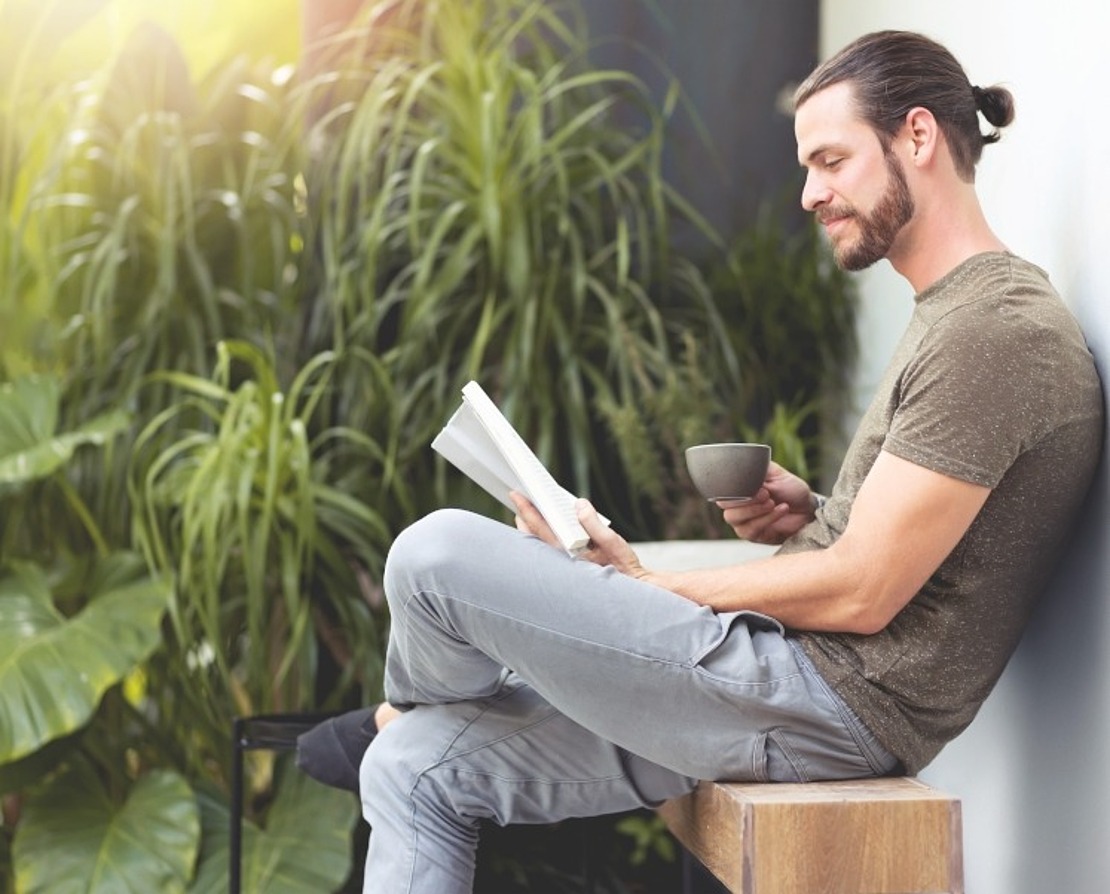 Ein Mann mit Bart und Pferdeschwanz sitzt auf einer Holzbank an einer Mauer, hält eine Tasse in der Hand und liest ein Buch. Er ist von üppigen grünen Pflanzen umgeben und wirkt im natürlichen Licht entspannt.
