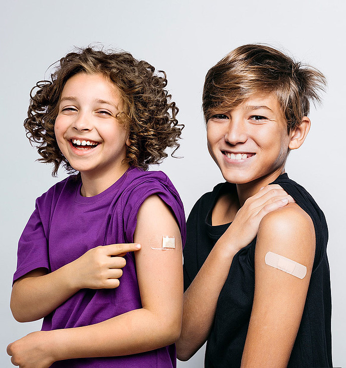 Zwei lächelnde Kinder stehen nebeneinander und zeigen Verbände an ihren Oberarmen, die darauf hinweisen, dass sie gerade geimpft worden sind. Ein Kind zeigt auf seinen Verband. Beide wirken glücklich und zuversichtlich vor einem schlichten Hintergrund.