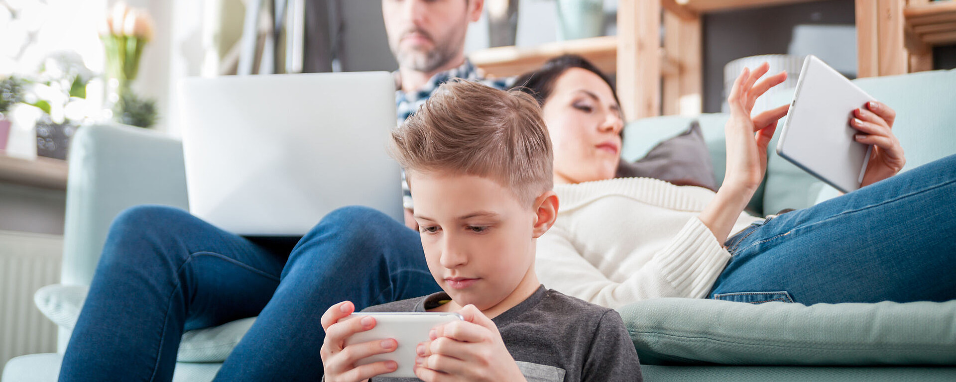 Ein Junge sitzt auf dem Boden und konzentriert sich auf sein Smartphone, während eine Frau mit einem Tablet auf der Couch liegt und ein Mann hinter ihr an einem Laptop arbeitet. Alle scheinen mit ihren Geräten in einem gemütlichen Wohnzimmer beschäftigt zu sein.