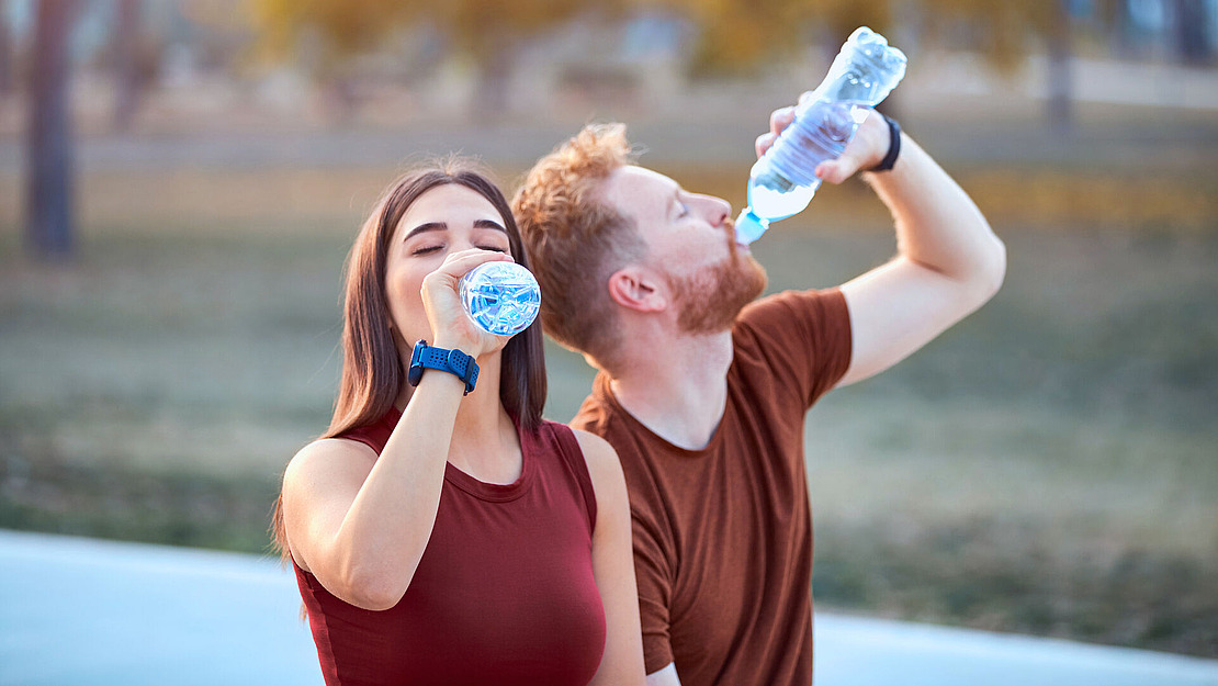 Eine Frau und ein Mann in legerer Sportkleidung trinken an einem sonnigen Tag im Freien Wasser aus Plastikflaschen, im Hintergrund sind verschwommene Bäume und Gras zu sehen.