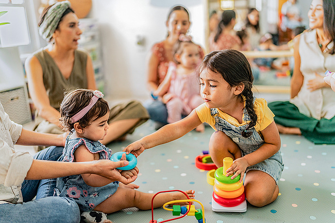 Mehrere kleine Kinder spielen mit buntem Spielzeug auf einer Spielmatte, während Erwachsene in der Nähe sitzen und sie beaufsichtigen. Ein Mädchen reicht einen Stapelring an ein Baby weiter, und alle scheinen in diesem hellen, fröhlichen Raum beschäftigt und konzentriert zu sein.