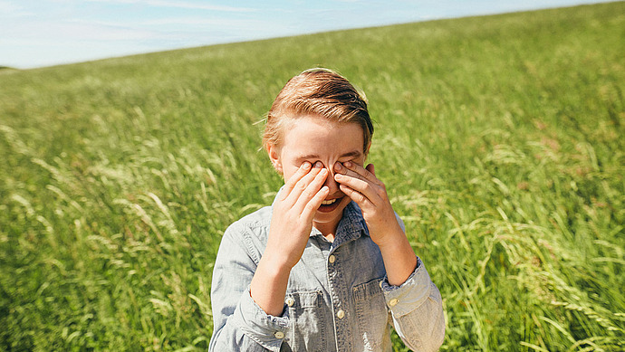 Ein kleines Kind in einem Jeanshemd steht lächelnd auf einer sonnigen grünen Wiese und bedeckt mit beiden Händen die Augen. Der Himmel ist klar und blau, und das Kind ist von hohem Gras umgeben.