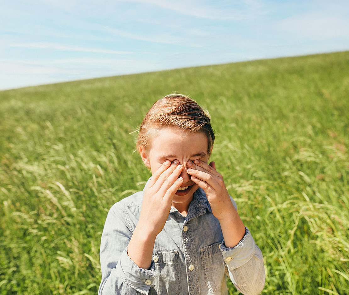Ein kleines Kind in einem Jeanshemd steht auf einer sonnigen grünen Wiese und bedeckt mit beiden Händen die Augen. Der Himmel ist klar und blau, und das Kind ist von hohem Gras umgeben.