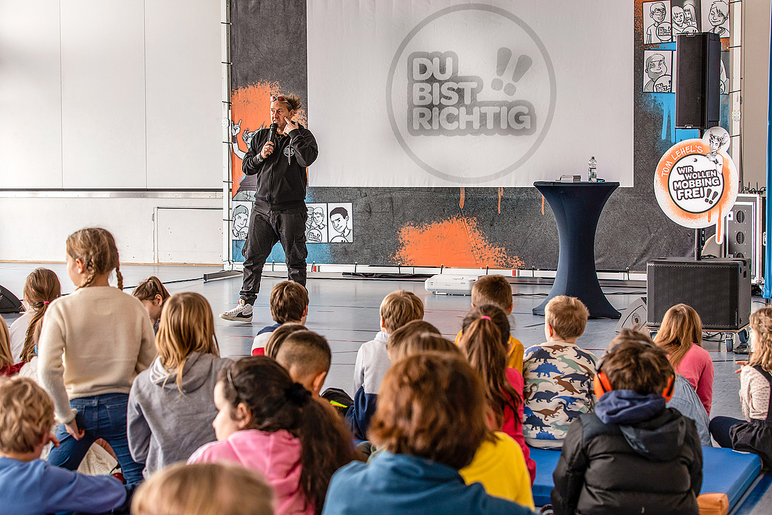 Ein Mann spricht in ein Mikrofon auf der Bühne vor einer Gruppe von Kindern, die auf dem Boden sitzen. Hinter ihm wird auf einer Leinwand "DU BIST RICHTIG" angezeigt, und um die Bühne herum sind bunte Dekorationen zu sehen.